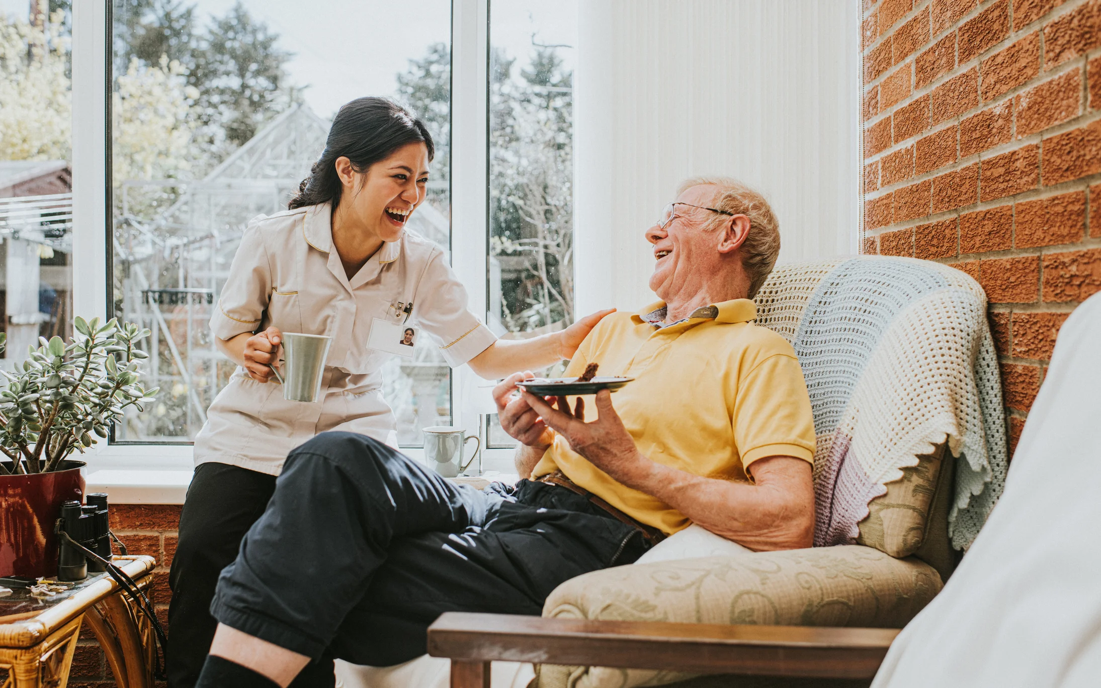 Assisted living caregiver laughing with a senior resident while serving tea in a bright sunroom